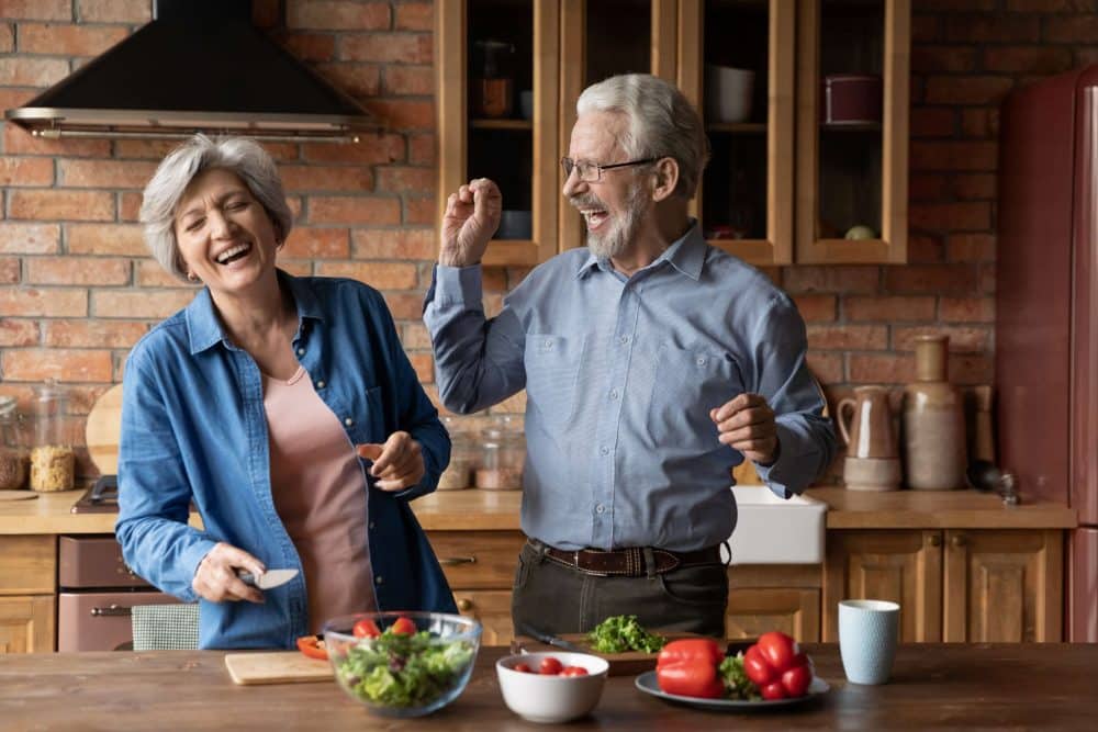 An older man and woman laugh and dance together while preparing a fresh vegetable salad at a rustic kitchen counter.